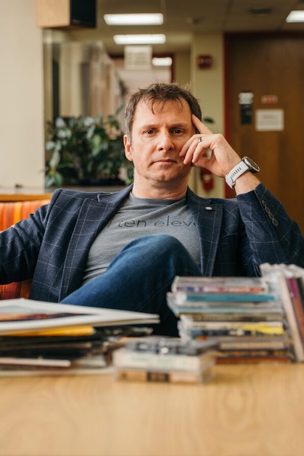 Headshot of faculty Director Jeremy Morris. He has short, brown hair and is wearing a gray undershirt, glue checkered jacket, blue jeans, and watch. He is sitting behind pile of vinyl records, cds, and cassette tapes.