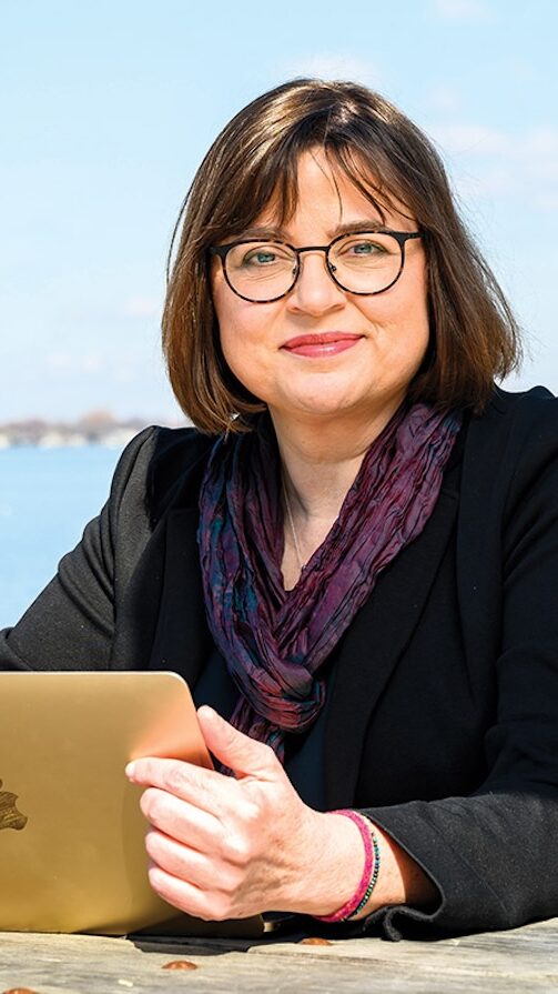 Headshot of Anja Wanner. She has shoulder length short brown hair, and is wearing glasses, a purple scarf, and a black jacket. She is sitting outside with an Apple laptop.