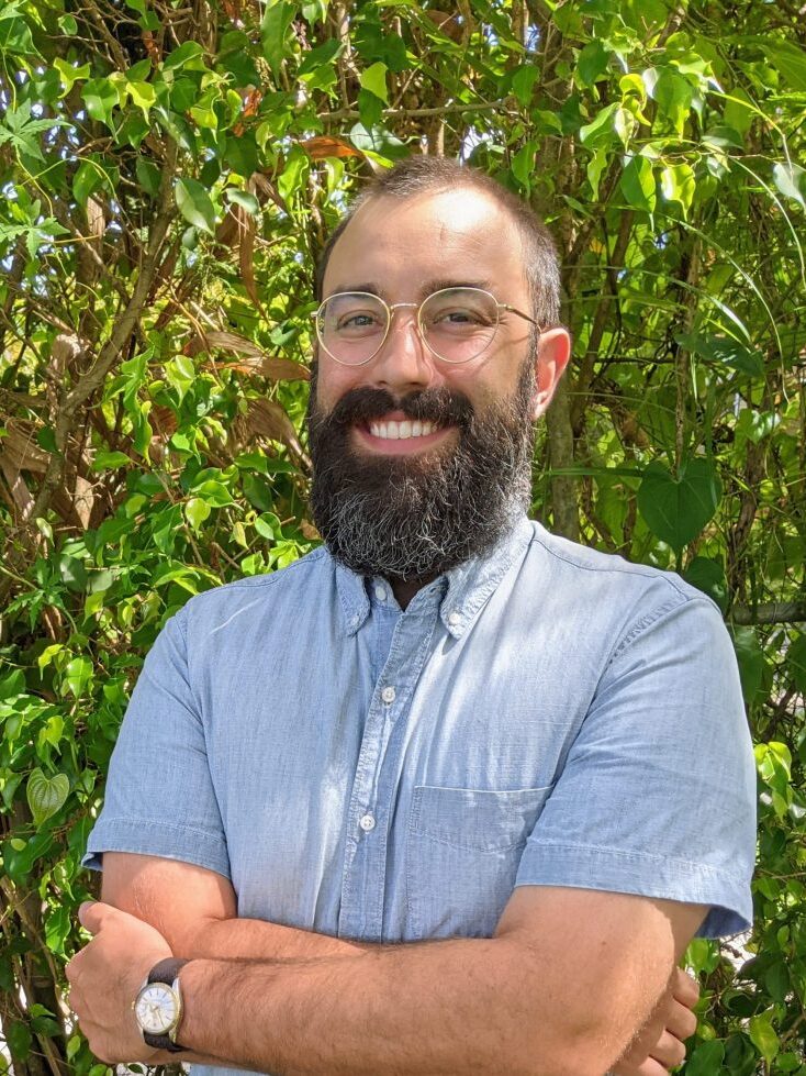 Headshot of Clinton Castro. He has short black hair, a beard, and is wearing glasses, a blue button up shirt, and a watch.