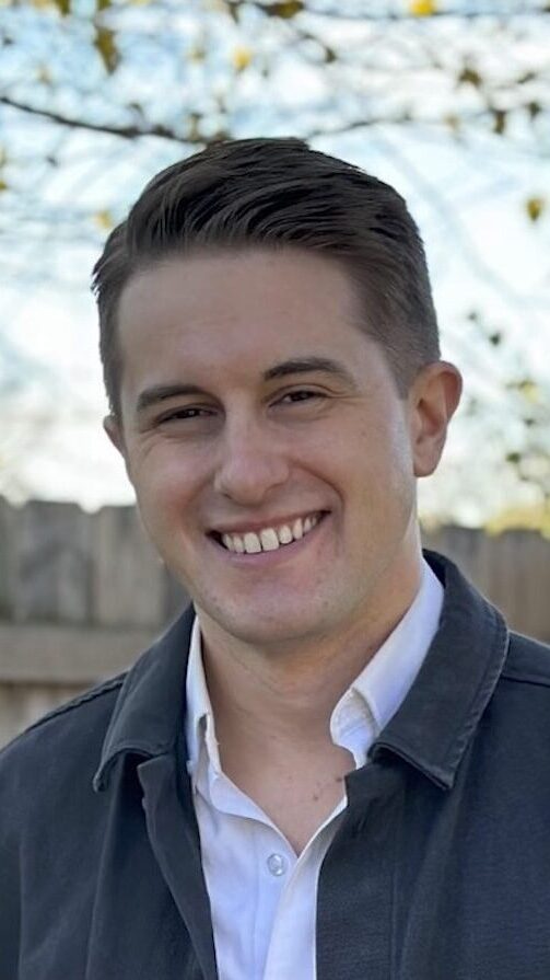 Headshot of Devin Kennedy. He has short, straight, brown hair, and is wearing a white button down shirt under a blue jacket.