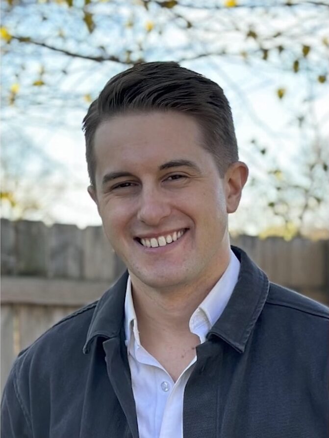 Headshot of Devin Kennedy. He has short, straight, brown hair, and is wearing a white button down shirt under a blue jacket.