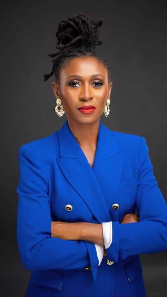 Headshot of Ainehi Edoro. She has curly brown hair styled in a bun, and is wearing gold earrings and a blue blazer.