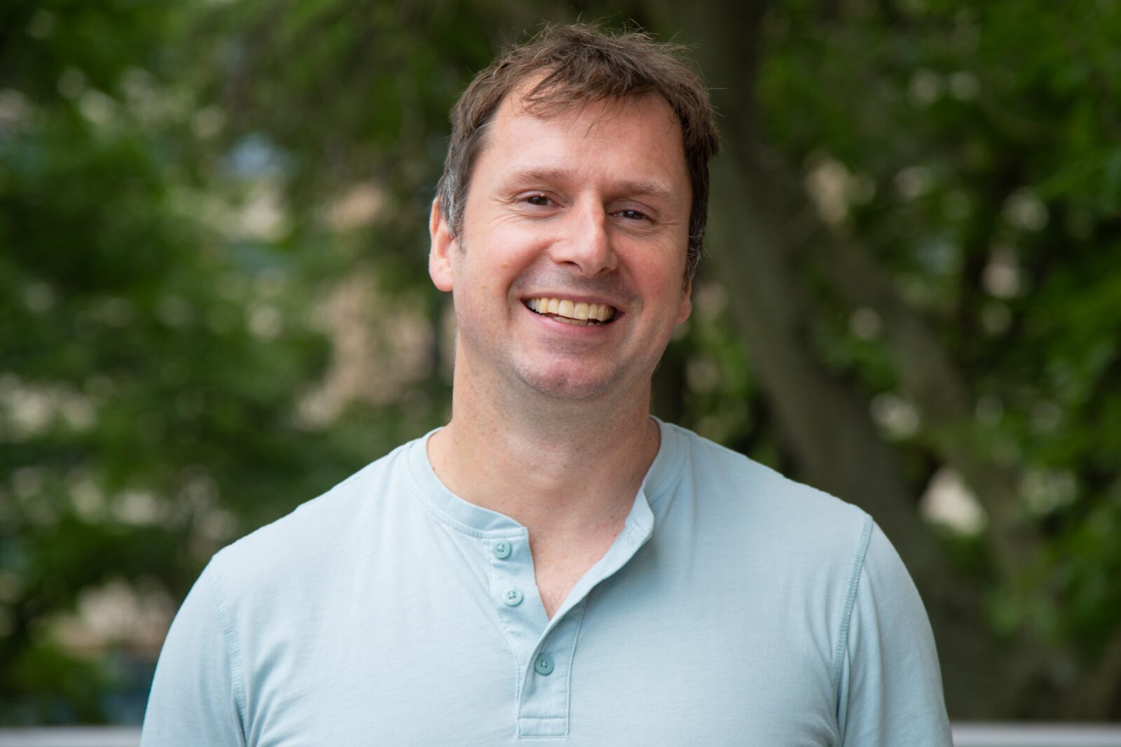 Picture of Faculty Director, Jeremy Morris, He has short brown hair and is wearing a blue shirt in front of a blurred out tree in the background