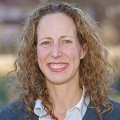 Photo of Susan Ridgley. She has long, curly, blond hair and is wearing a white button down shirt under a blue/gray sweater. 