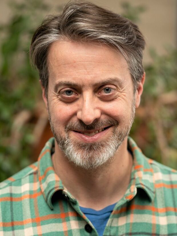 Headshot of Alan Rubel. He has short hair with brown and gray, and is wearing a green and oranged checkered collared shirt over a blue undershirt.
